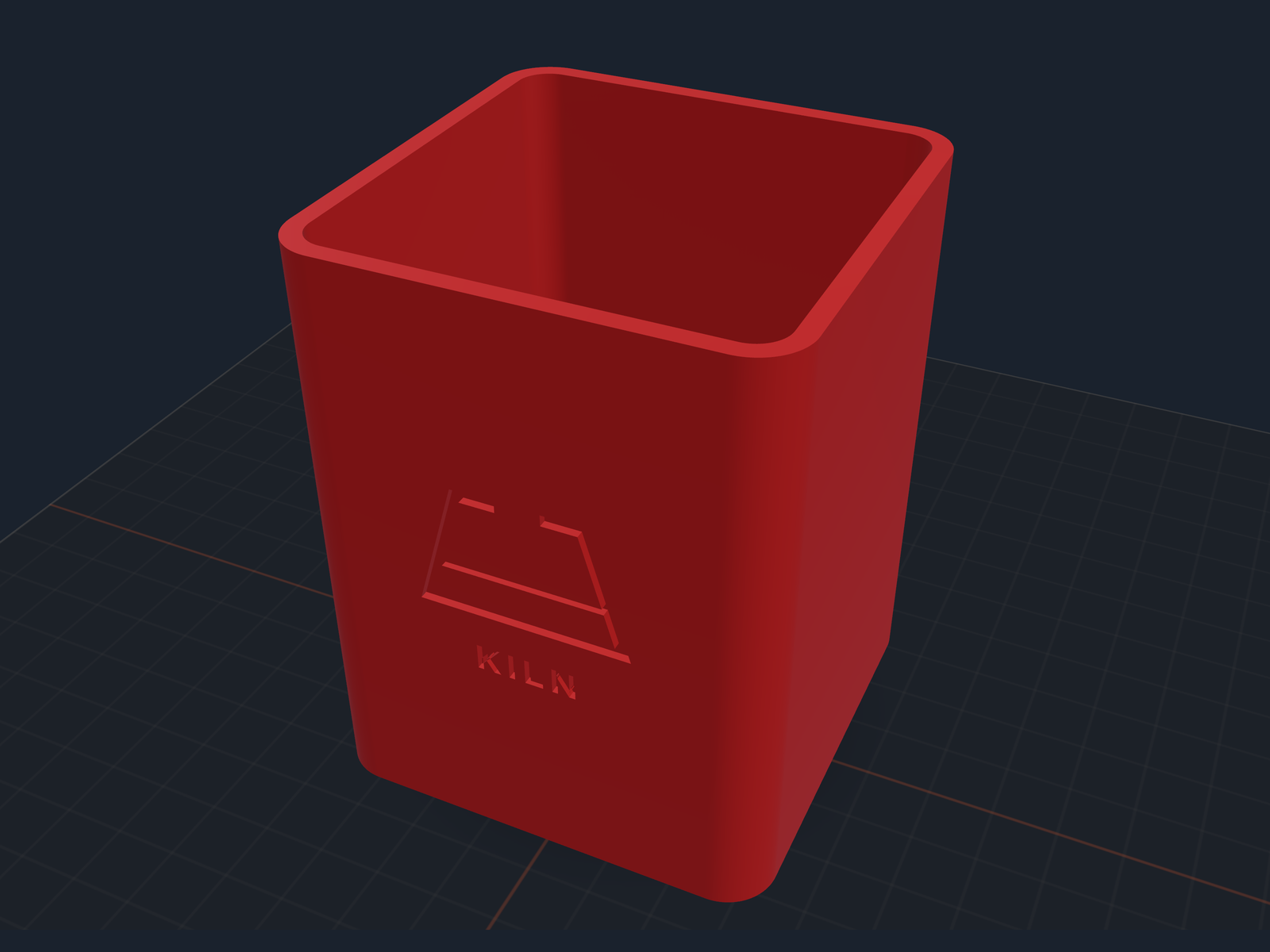 A red pen cup with the Kiln mark and the word KILN carved into its front face, lit from the side on a dark print bed with a soft shadow beneath it