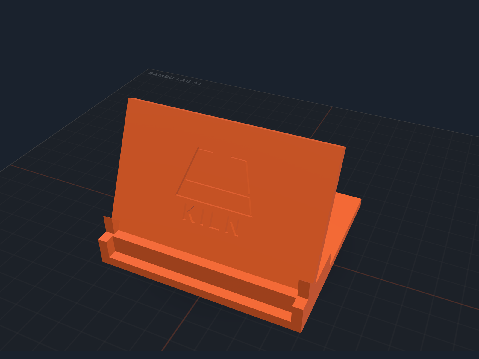An orange printed phone stand on a dark print bed, with the Kiln mark and the word KILN cut into its leaning back panel and the surface around them left uncut